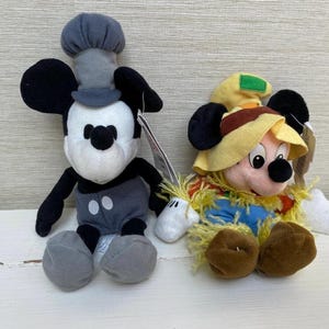 May include: Two Mickey Mouse soft toys. One is dressed as a chef in grey and black, the other as a scarecrow in a straw hat and blue overalls. Both have black ears and are on a white surface.