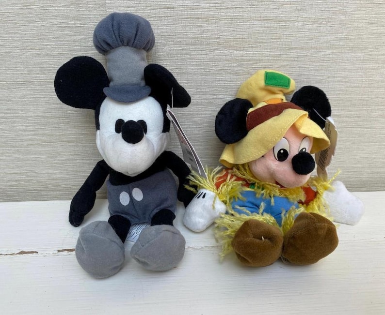 May include: Two Mickey Mouse soft toys. One is dressed as a chef in grey and black, the other as a scarecrow in a straw hat and blue overalls. Both have black ears and are on a white surface.