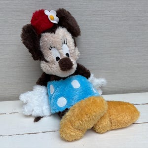 May include: A plush Minnie Mouse doll with a red bow and white flower on her head. She has a tan face, brown ears, and a blue skirt with white polka dots. Her legs are yellow and her hands are white.