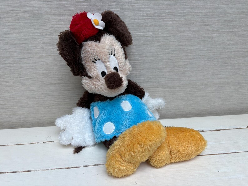 May include: A plush Minnie Mouse doll with a red bow and white flower on her head. She has a tan face, brown ears, and a blue skirt with white polka dots. Her legs are yellow and her hands are white.