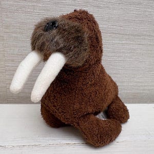 May include: A brown and tan plush walrus toy. The soft toy features large white tusks and a furry face. The walrus is sitting upright, with its flippers and body visible.