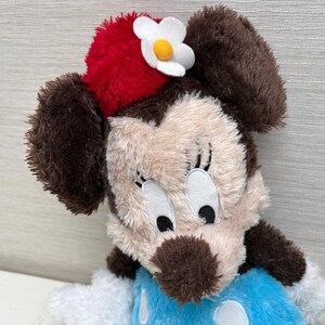 May include: A plush Minnie Mouse doll with brown ears, a red hat with a white flower, and a blue dress with white polka dots. The doll has a beige face with black eyes and a brown nose.