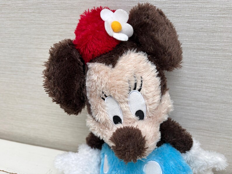 May include: A plush Minnie Mouse doll with brown ears, a red hat with a white flower, and a blue dress with white polka dots. The doll has a beige face with black eyes and a brown nose.