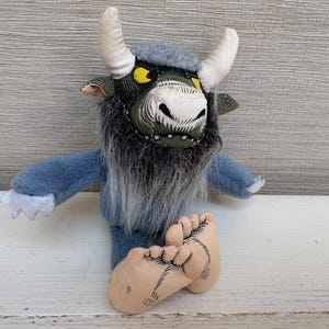 May include: A plush monster toy featuring a blue body, long gray faux fur chest, and two white horns. The creature has a green patterned face with yellow eyes, a white snout, and large beige feet with black line details on the toes and soles.