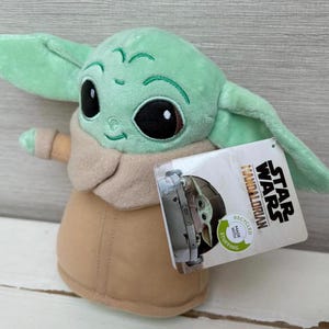 Peut inclure: Peluche de Grogu, &eacute;galement connu sous le nom de Baby Yoda, de Star Wars: The Mandalorian. Le jouet est vert clair avec de grands yeux noirs, des v&ecirc;tements beiges et de grandes oreilles. Une &eacute;tiquette Star Wars est attach&eacute;e.