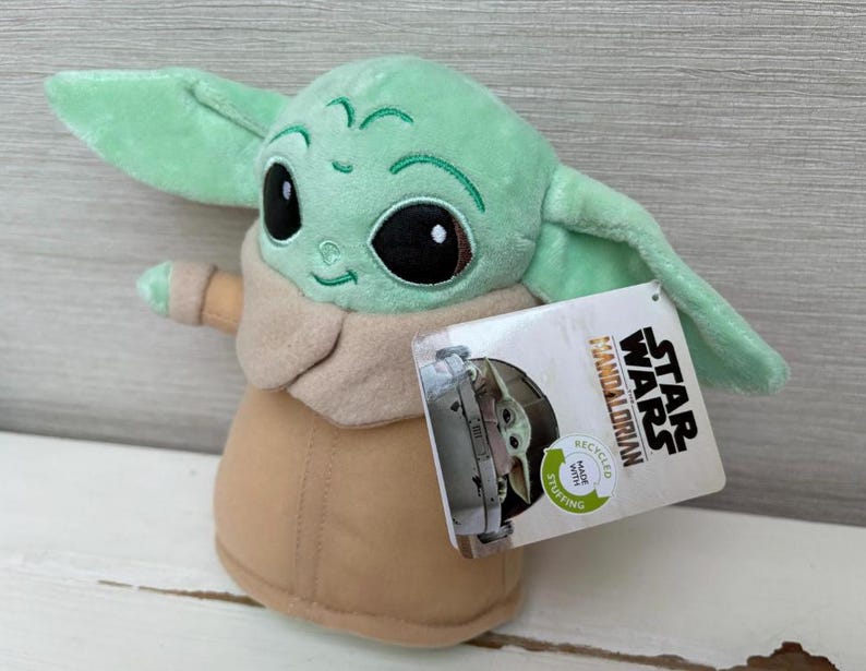 Peut inclure: Peluche de Grogu, &eacute;galement connu sous le nom de Baby Yoda, de Star Wars: The Mandalorian. Le jouet est vert clair avec de grands yeux noirs, des v&ecirc;tements beiges et de grandes oreilles. Une &eacute;tiquette Star Wars est attach&eacute;e.