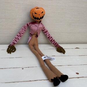 May include: A plush doll featuring a round orange pumpkin head with a stitched black jagged mouth and eyes. The doll has a long-sleeved pink velvet jacket, tan ribbed fabric legs, dark brown hands, and black feet with a white tag attached.