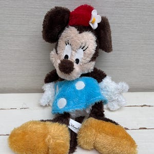 May include: A plush Minnie Mouse doll with brown fur, a red bow, and a blue dress with white polka dots. The doll has large, round ears and yellow feet. The Disney Parks tag is visible.