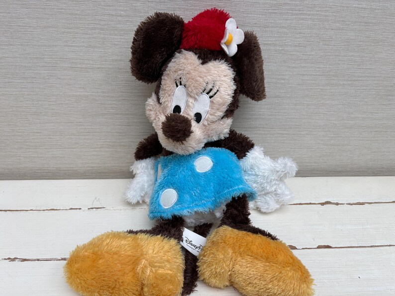 May include: A plush Minnie Mouse doll with brown fur, a red bow, and a blue dress with white polka dots. The doll has large, round ears and yellow feet. The Disney Parks tag is visible.