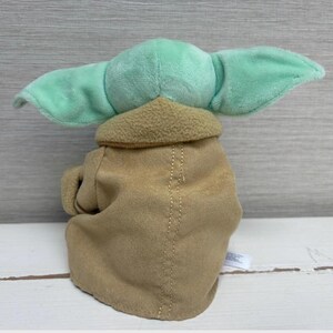 May include: A soft toy of a character with large, mint green ears and a tan robe. The toy has a light brown collar and a stitched seam down the centre of the robe. The toy is a popular character from a science fiction franchise.