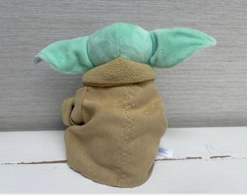 May include: A soft toy of a character with large, mint green ears and a tan robe. The toy has a light brown collar and a stitched seam down the centre of the robe. The toy is a popular character from a science fiction franchise.