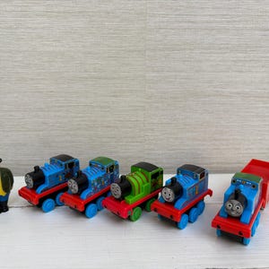 May include: A set of toy train engines and a figure. The trains are blue, green, and red, with smiling faces. The figure is dressed in a green coat, yellow shirt, blue trousers, and a black hat. The toys are on a white surface.