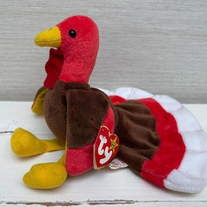 Peut inclure: Une peluche de dinde avec une tête et un fanon rouges, un corps marron et une queue en éventail de plumes rouges et blanches. La dinde a des pieds jaunes et un bec jaune. Une étiquette rouge en forme de cœur est attachée au jouet.