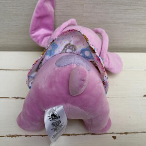 Puede incluir: Un peluche de color lavanda con orejas grandes y una cola. El peluche tiene una tela estampada en la espalda con rosquillas y otras golosinas. Se ve una etiqueta de Disney.