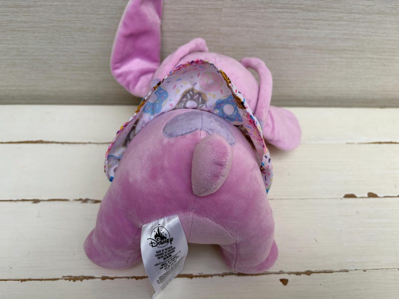 Puede incluir: Un peluche de color lavanda con orejas grandes y una cola. El peluche tiene una tela estampada en la espalda con rosquillas y otras golosinas. Se ve una etiqueta de Disney.
