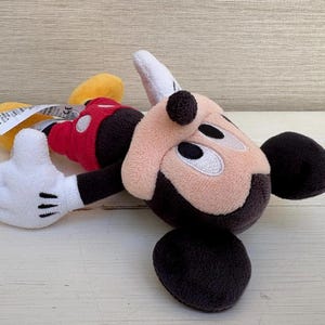 K&ouml;nnte beinhalten: Ein Stofftier von Micky Maus in liegender Position. Das Spielzeug hat ein beiges Gesicht, schwarze Ohren und eine schwarze Nase. Es tr&auml;gt eine rote Weste, schwarze Shorts, gelbe Schuhe und wei&szlig;e Handschuhe mit schwarzen Linien.