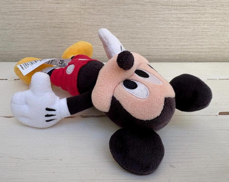 K&ouml;nnte beinhalten: Ein Stofftier von Micky Maus in liegender Position. Das Spielzeug hat ein beiges Gesicht, schwarze Ohren und eine schwarze Nase. Es tr&auml;gt eine rote Weste, schwarze Shorts, gelbe Schuhe und wei&szlig;e Handschuhe mit schwarzen Linien.