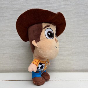 May include: A plush toy of a cartoon cowboy character, featuring a brown cowboy hat, tan face, and a yellow and black plaid shirt. The toy has blue pants and brown boots, with large, expressive eyes.