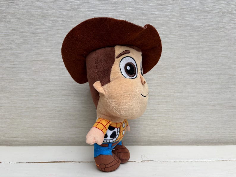 May include: A plush toy of a cartoon cowboy character, featuring a brown cowboy hat, tan face, and a yellow and black plaid shirt. The toy has blue pants and brown boots, with large, expressive eyes.