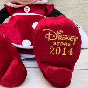 May include: A plush Minnie Mouse doll in a red dress with white trim and buttons. The doll's foot has the text "Disney Store 2014" in gold lettering. The doll has black arms and white gloves.