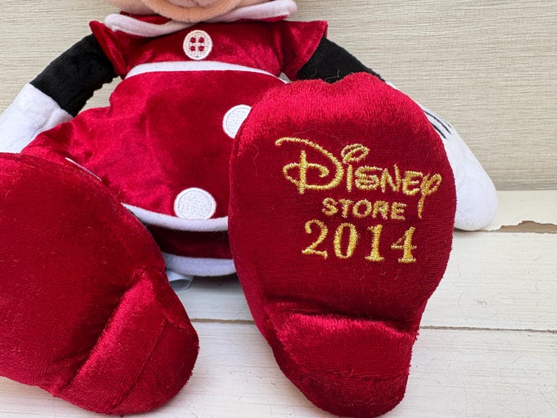 May include: A plush Minnie Mouse doll in a red dress with white trim and buttons. The doll's foot has the text "Disney Store 2014" in gold lettering. The doll has black arms and white gloves.