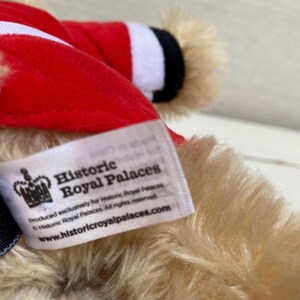 May include: A plush, tan teddy bear wearing a red and white striped outfit. The bear has a tag that reads "Historic Royal Palaces" and the website address. The bear's fur is soft and fluffy.