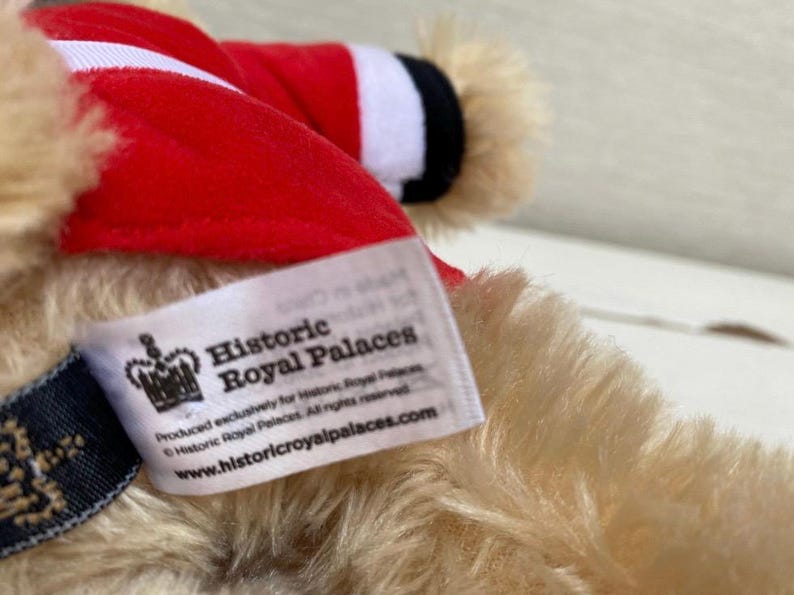 May include: A plush, tan teddy bear wearing a red and white striped outfit. The bear has a tag that reads "Historic Royal Palaces" and the website address. The bear's fur is soft and fluffy.