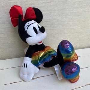 May include: A plush Minnie Mouse doll in a relaxed pose. The doll has a black body, white face and hands, and a red bow. It wears a rainbow-coloured skirt and has rainbow-coloured accents on its feet.