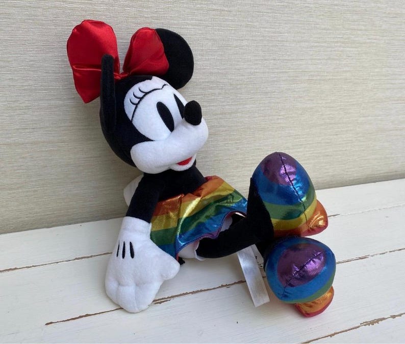 May include: A plush Minnie Mouse doll in a relaxed pose. The doll has a black body, white face and hands, and a red bow. It wears a rainbow-coloured skirt and has rainbow-coloured accents on its feet.