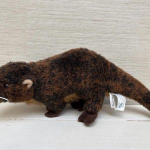 Puede incluir: Un juguete de peluche de nutria marrón con un vientre marrón claro y pequeñas patas marrones oscuras. El animal de peluche tiene un cuerpo largo, una cabeza pequeña con un hocico puntiagudo y bigotes blancos. Una etiqueta está adherida al juguete.