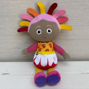 Può includere: Bambola di peluche con viso marrone, grandi occhi bianchi e espressione sorridente. La bambola ha una testa rosa con accenti colorati a forma di petalo. Indossa un top floreale giallo e rosso, una gonna bianca con rifiniture rosse e scarpe rosa e viola.
