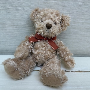 May include: A soft teddy bear with light brown fur and a dark brown nose. The bear has a red ribbon tied in a bow around its neck. The bear is sitting on a light grey wooden surface.