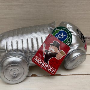 May include: A silver-colored plush toy shaped like a classic race car token from the Monopoly board game. The toy features stitched detailing, a red tag with an illustration of the Monopoly mascot, and a green circular tag indicating recycled filling.
