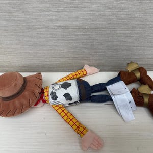 May include: A plush toy of a cowboy character, featuring a brown hat, yellow and red plaid sleeves, a cow-print vest, blue jeans, and brown boots with gold accents. The toy is lying on a white surface.