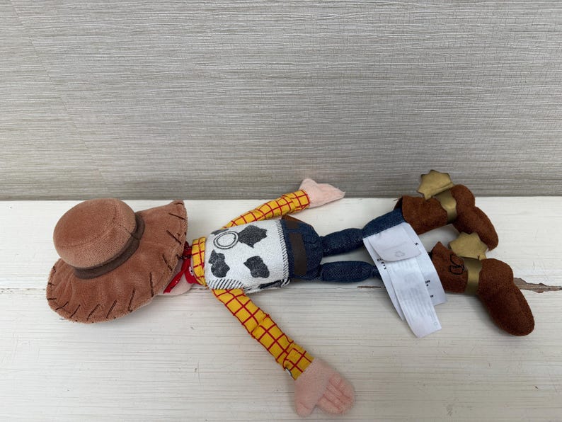 May include: A plush toy of a cowboy character, featuring a brown hat, yellow and red plaid sleeves, a cow-print vest, blue jeans, and brown boots with gold accents. The toy is lying on a white surface.