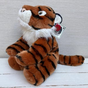 May include: A plush tiger toy with brown and black stripes, white fur around the neck, and a tag attached. The toy is sitting on a white surface. The tiger's fur is a mix of brown and orange, with black stripes.