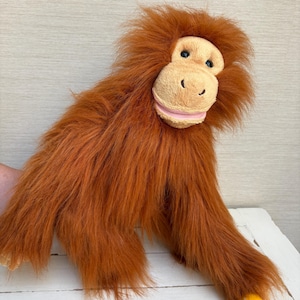 Op de afbeelding: Een orang-oetan knuffel met lang, ruig, feloranje bont, een lichtbruin gezicht met donkere ogen en een zachte roze mond. Het knuffeldier heeft lange armen en een enkel zichtbaar geel en wit voetdetail in de rechterbenedenhoek.