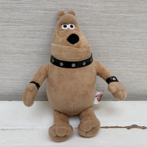 Puede incluir: Un juguete de peluche con forma de perro de color tostado, con un collar negro con tachuelas plateadas y muñequeras negras a juego. El peluche tiene un cuerpo redondeado, orejas pequeñas, una nariz negra y ojos blancos con pupilas negras.
