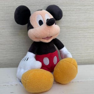 K&ouml;nnte beinhalten: Ein Pl&uuml;schtier von Mickey Mouse mit schwarzen Ohren, einem schwarzen K&ouml;rper, roten Shorts und gelben F&uuml;&szlig;en. Das Spielzeug hat wei&szlig;e Handschuhe und ein l&auml;chelndes Gesicht mit einem rosa Mund. Der Hintergrund ist eine helle Holzmaserung.