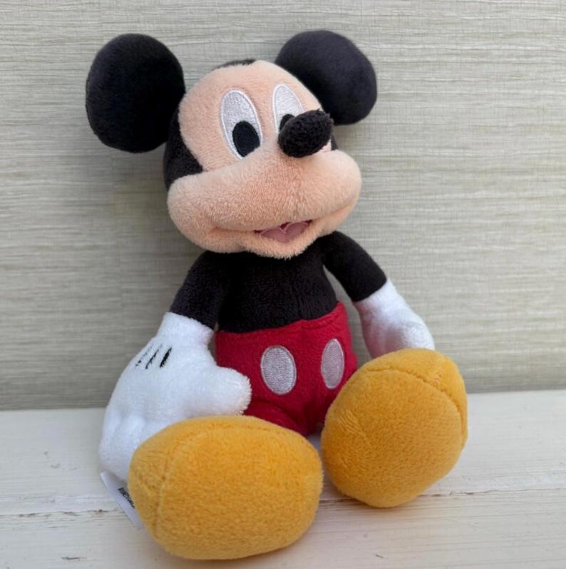 K&ouml;nnte beinhalten: Ein Pl&uuml;schtier von Mickey Mouse mit schwarzen Ohren, einem schwarzen K&ouml;rper, roten Shorts und gelben F&uuml;&szlig;en. Das Spielzeug hat wei&szlig;e Handschuhe und ein l&auml;chelndes Gesicht mit einem rosa Mund. Der Hintergrund ist eine helle Holzmaserung.