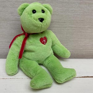 May include: A green plush teddy bear with a red ribbon around its neck and a red heart-shaped emblem on its chest. The bear has black eyes and a black nose. The bear is sitting on a white wooden surface.