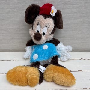May include: A plush Minnie Mouse doll with brown fur, a red bow, and a blue dress with white polka dots. The doll has large, round ears and oversized yellow feet. A Disney Parks tag is attached.