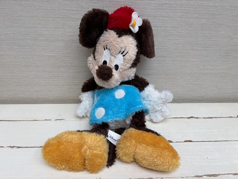 May include: A plush Minnie Mouse doll with brown fur, a red bow, and a blue dress with white polka dots. The doll has large, round ears and oversized yellow feet. A Disney Parks tag is attached.