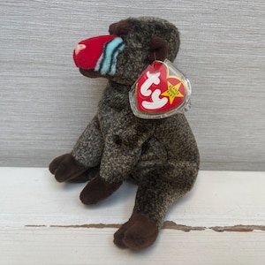 May include: A plush toy baboon with a red nose, blue stripes, and brown fur. The baboon has dark brown feet and a red and yellow tag attached to its ear. The toy is sitting on a white surface.