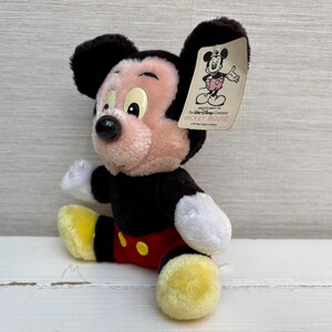 Puede incluir: Un peluche de Mickey Mouse sentado. El juguete tiene orejas negras, cuerpo negro y cara rosa. Lleva pantalones cortos rojos, zapatos amarillos y guantes blancos. Una etiqueta con un gr&aacute;fico de Mickey Mouse est&aacute; adjunta.