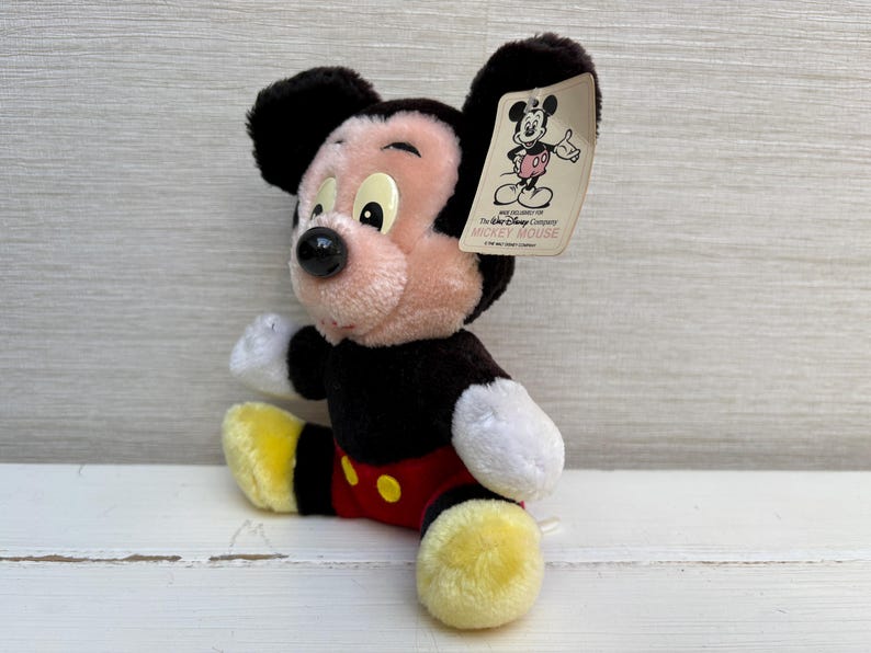 Puede incluir: Un peluche de Mickey Mouse sentado. El juguete tiene orejas negras, cuerpo negro y cara rosa. Lleva pantalones cortos rojos, zapatos amarillos y guantes blancos. Una etiqueta con un gr&aacute;fico de Mickey Mouse est&aacute; adjunta.