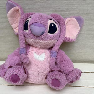 May include: A plush toy of the character Angel from the Disney movie Lilo & Stitch. The toy is primarily purple with pink accents on the ears, chest, and feet. It has large black eyes and a heart-shaped design on its chest.