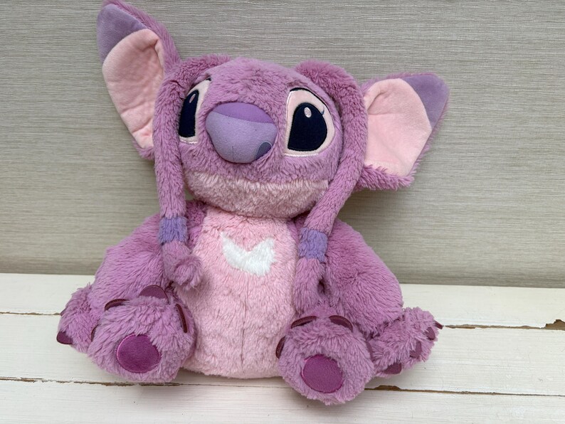 May include: A plush toy of the character Angel from the Disney movie Lilo & Stitch. The toy is primarily purple with pink accents on the ears, chest, and feet. It has large black eyes and a heart-shaped design on its chest.