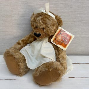 May include: A brown plush teddy bear with shaggy fur, wearing a cream-colored fabric dress and a matching ribbon bow on its head. A square paper tag attached to the bear features an illustration of a bear and the text Alice's Bear Shop.
