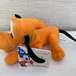 Pode incluir: Pelúcia Pluto laranja com orelhas e cauda pretas, uma coleira verde e uma língua rosa. A pelúcia tem uma etiqueta com Mickey Mouse com um chapéu de mago. A etiqueta diz "Disneyland Paris" e "The World of Plushies."